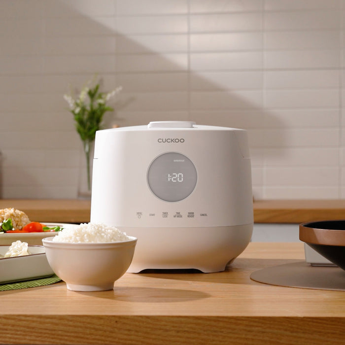 White Cuckoo rice cooker on a wooden kitchen counter, with a bowl of cooked white rice in the foreground. (CR-0661F)
