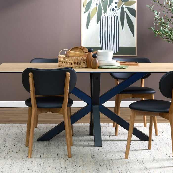 A modern dining table set in a styled room, featuring a rectangular light wood table with a black X-shaped base, surrounded by four chairs with black seats and light wood frames. A white and green botanical art print hangs on the wall behind the table.