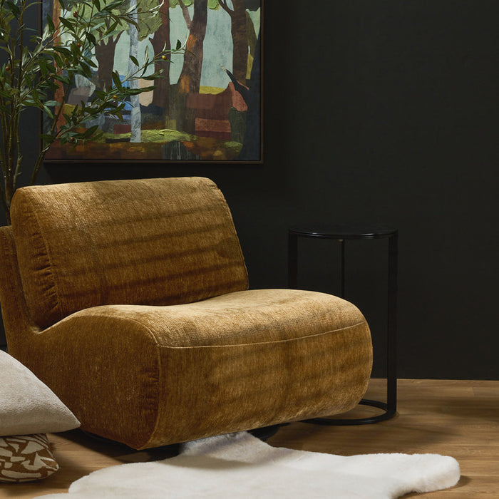 A modern mustard yellow velvet armchair in a styled living room setting with a dark charcoal wall, a framed forest painting, a small black side table, a white cowhide rug, and a potted plant.