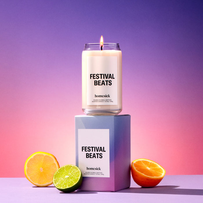 A lit Festival Beats Homesick candle displayed on top of its boxed packaging on a colorful background and surrounded by halved citrus fruits.