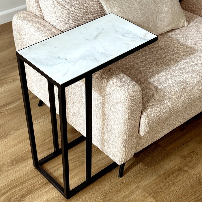 A modern side table with a white marble-patterned top and a black metal frame, positioned next to a beige textured sofa on a light wood floor.