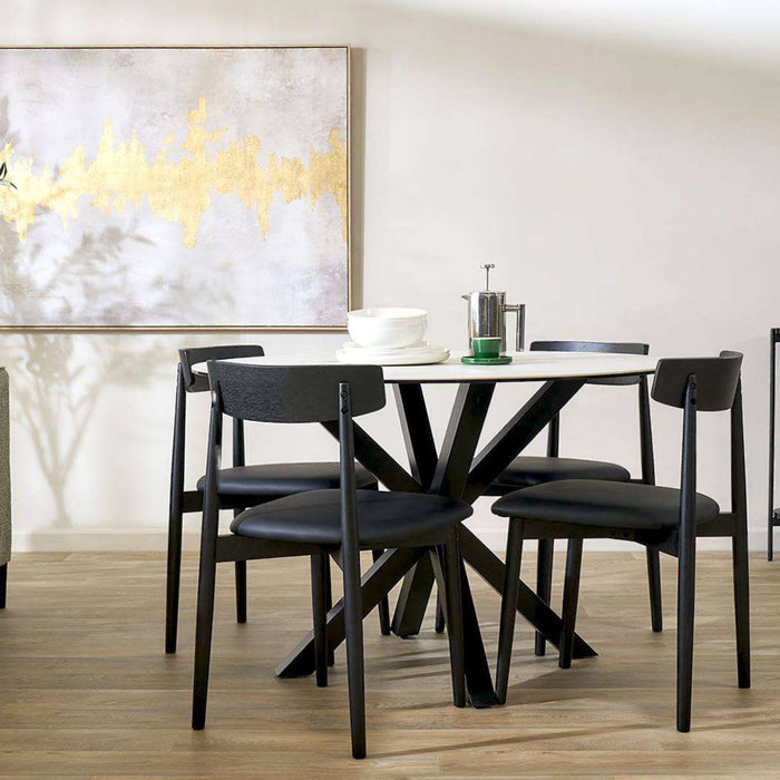 A modern dining table set with four black chairs in a bright dining room. The table has a white top and a black X-shaped pedestal base, with a white bowl and a green-topped metal pitcher on top.