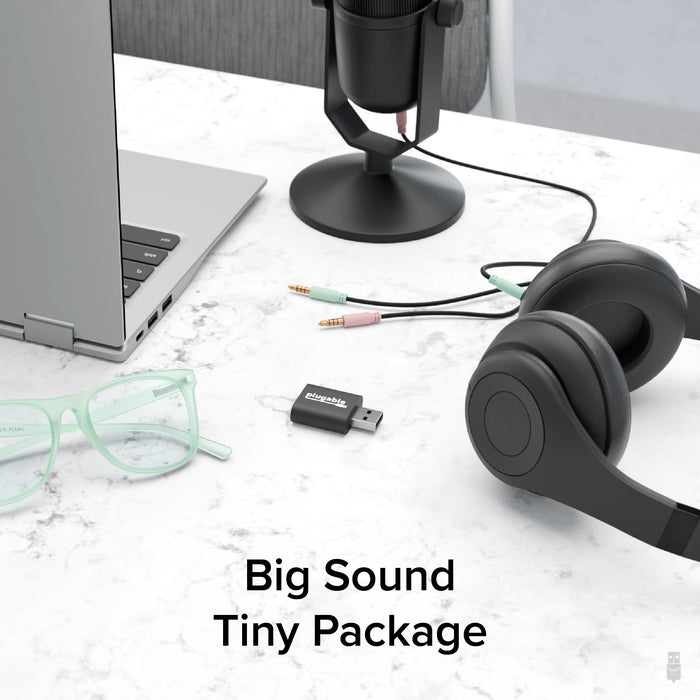 Plugable USB-AUDIO adapter on a desk with a laptop, microphone, and headphones, illustrating its compact form factor.