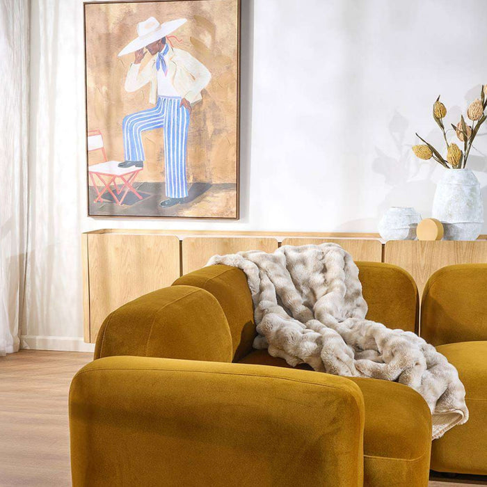 A mustard yellow velvet armchair in a bright, modern living room, styled with a chunky light gray knit throw blanket draped over the seat. A light wood sideboard with a colorful painting hangs on the white wall behind it.