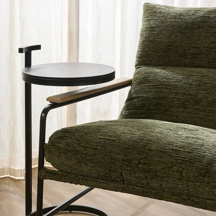 A modern side table with a round black top, black metal frame, and a light wood armrest, shown next to an olive green armchair in a room with sheer curtains and a wooden floor.