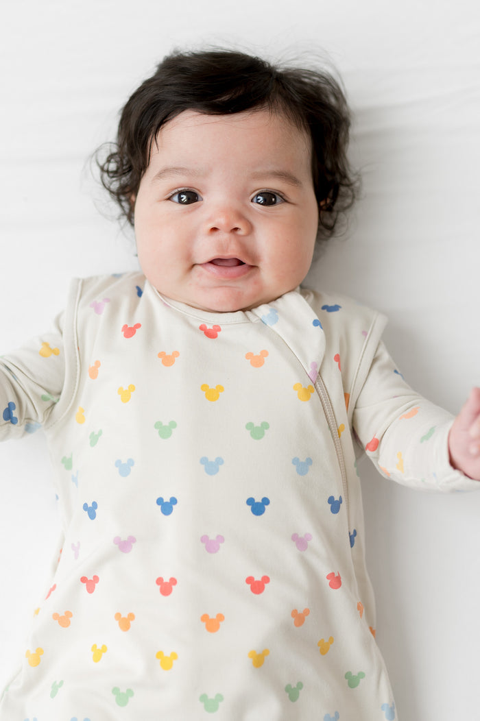 Baby wearing a cream sleep sack with colorful mickey patterns on a white background
