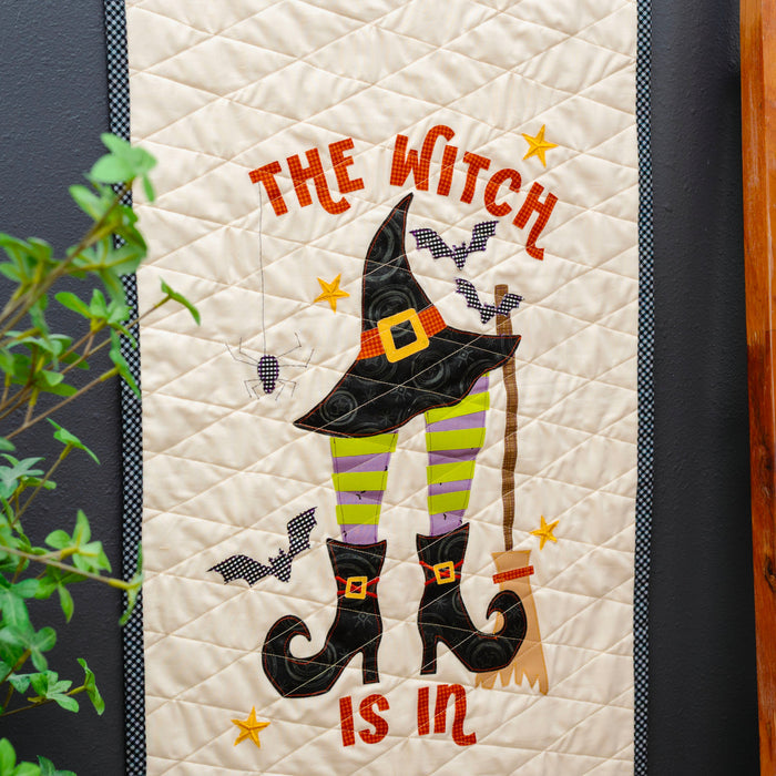 A vertical quilted wall hanging or tapestry with a Halloween witch design, shown hanging on a dark wall next to a wooden ladder holding another quilt. The main design features the words 'THE WITCH IS IN' in orange, a black witch hat, green-and-yellow striped legs, black boots, bats, and stars.