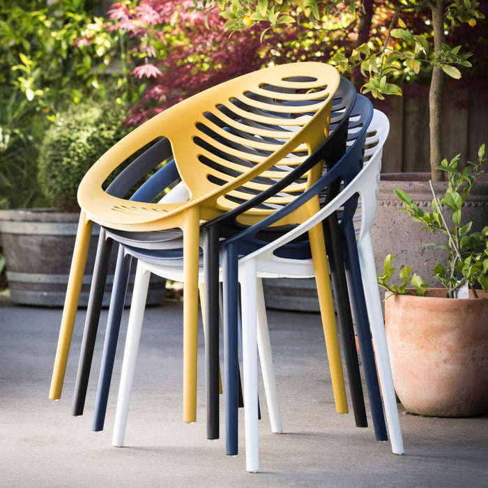 Three modern molded outdoor chairs in yellow, navy blue, and white, stacked and leaning against a garden patio background with potted plants.
