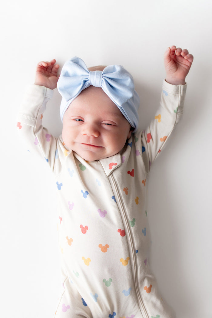 Baby wearing a colorful mickey footed onesie with mickey head prints and a light blue bow headband on a white background.